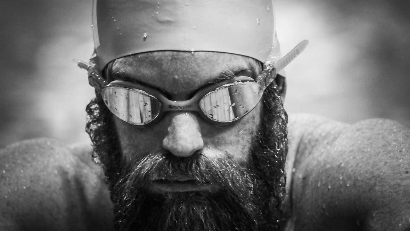 Close up headshot of man with a beard wearing swimming goggles and hat, covered in water droplets.