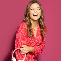 A laughing woman in a red dress holding a pair of overear headphones against plain dark pink background.