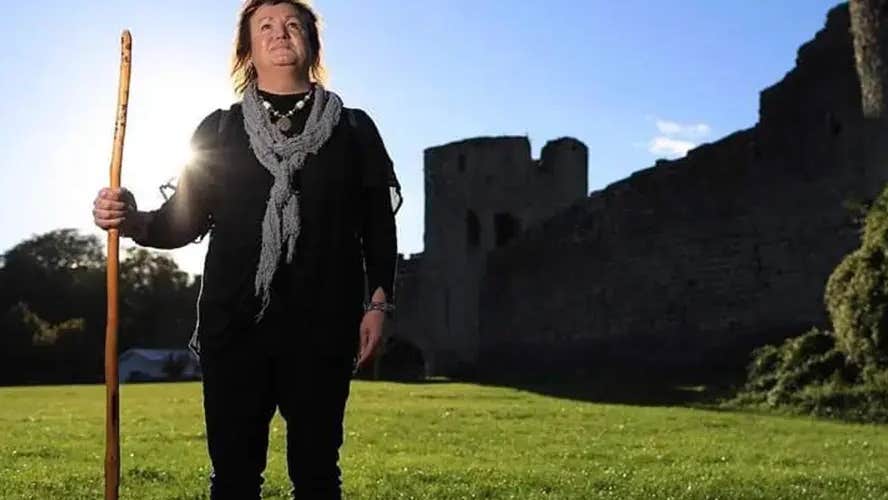 A person standing in a field holding a tall stick against pale blue sky with outline of turret and walls.