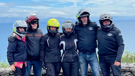 A group of bikers with their helmets on standing side by side at a coastal view