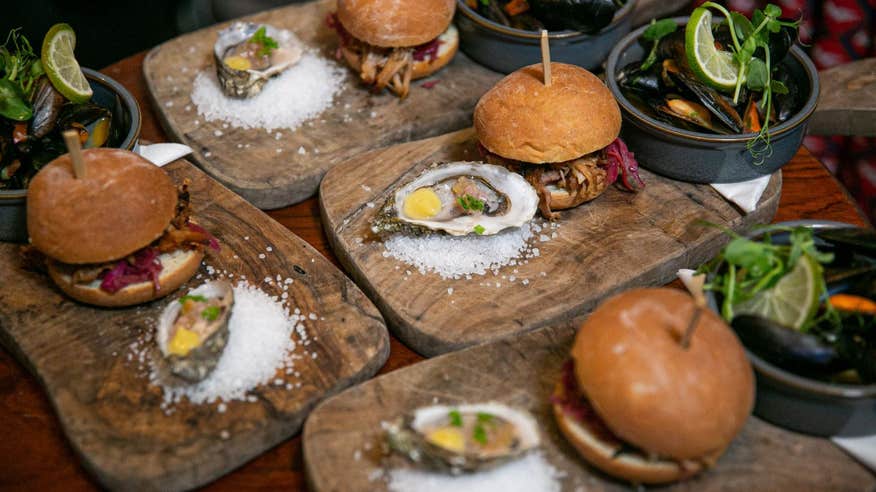 Four burgers on lush buns with a wooden stick placed in the middle to keep them standing upright all on different bread boards with an oyster on the side as well as a side salad.