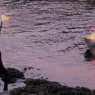 A person in a cloak holds a lit torch, watching a pire blaze in the middle of a misty river.