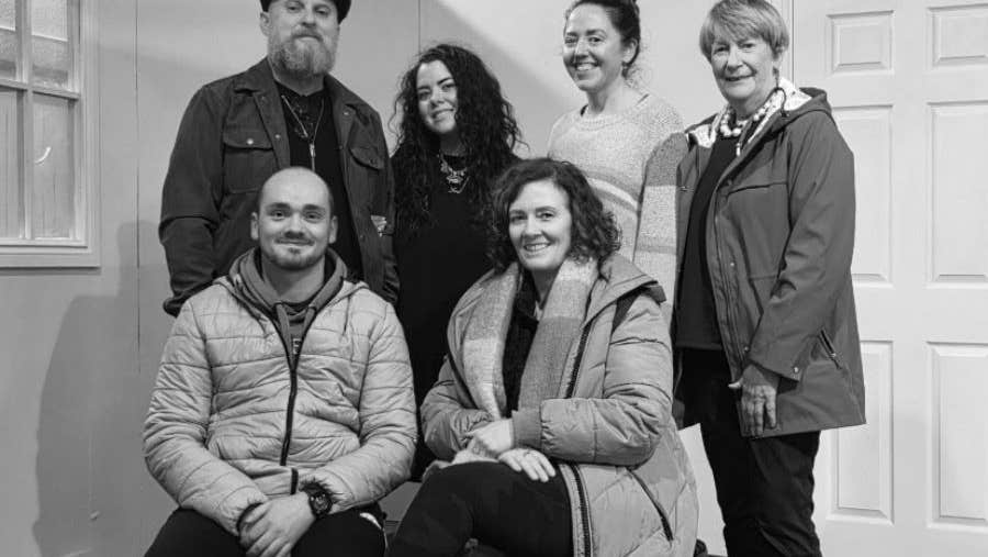 A group of smiling people with 2 seated, the rested standing, some wearing coats indoors.