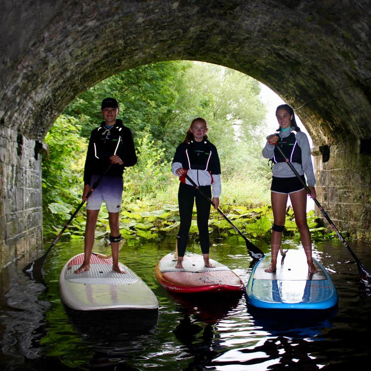 Three people in shorts standing on stand up paddle boards in water holding oars beneath a tunnel