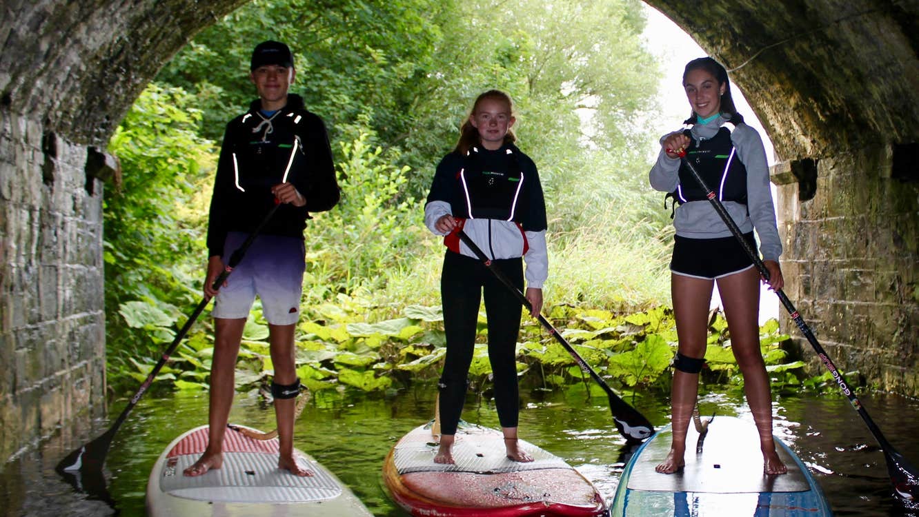 Three people in shorts standing on stand up paddle boards in water holding oars beneath a tunnel