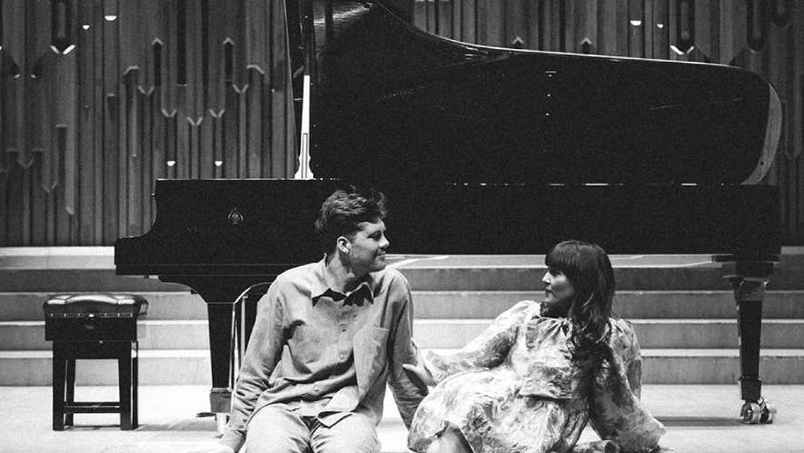 Black and white photo of 2 people seated on front edge of a stage looking directly at each other with a grand piano and stool behind them.