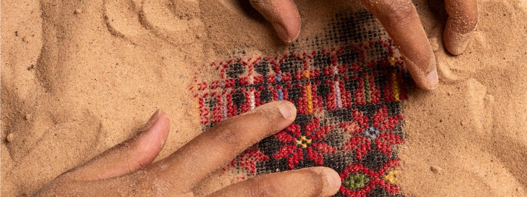 Looking down at a pair of hands digging out a small square of red and black patterned cloth from sand.