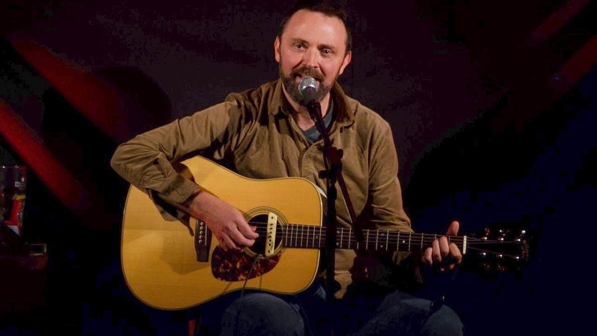 A man seated playing a guitar, singing into a mic on a stand