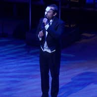 A man in evening dress singing into a mic on a dark stage, half of his face covered with a small mask.