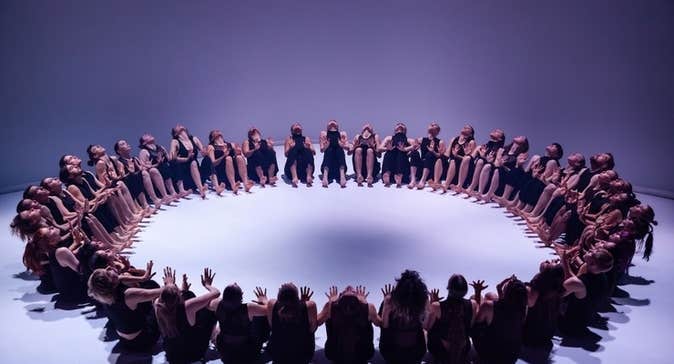 A large group of dancers seated on a white floor in a large circle.