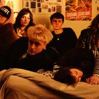 A group of 6 people seated on a bed in a room with poster on the wall behind
