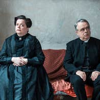 A serious looking man and woman dressed in black seated on a large sofa