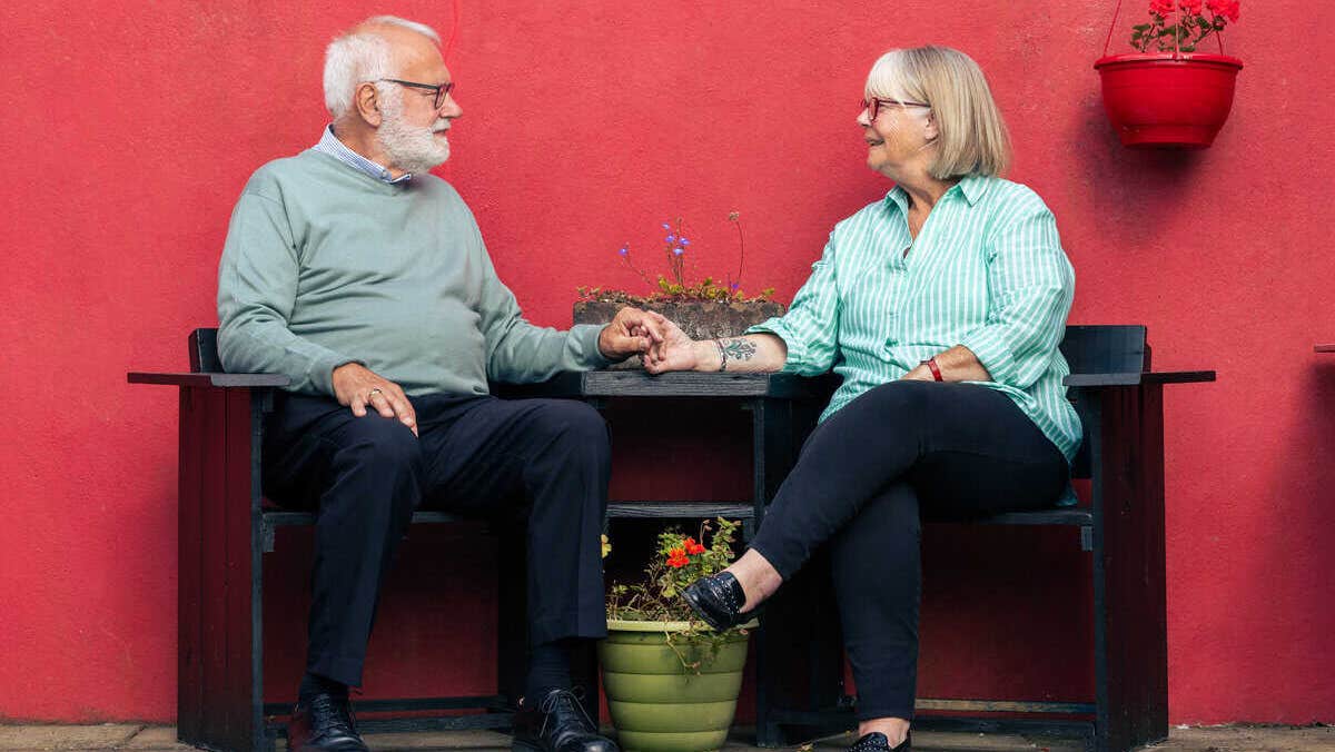 A man and woman seated on dark seats looking at each other, holding hands.