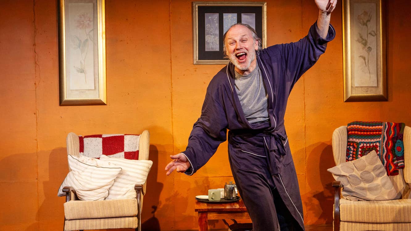 An animated man in grey dressing gown with arms outstretched with arms chairs and table in the background on a stage set.