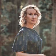 Slightly side profile on woman in dark, patterned dress with blurry trees in background.