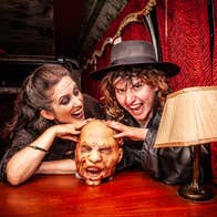 Two characters in costume with a disembodied model head on a table