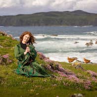 Person dressed in green tartan sitting among pink wildflowers on a grassy hillside near the sea