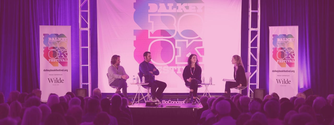 Fuzzy, pink lit view of a stage with 4 seated people in a discussion with a seated audience.