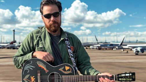 A bearded man in sunglasses is holding an ornately decorated black guitar with an airport and planes in the background.