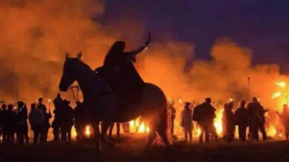 Person on a horse and line of figures at night silhouetted against flames and red smoke.
