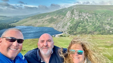 A couple and a tour guide posing for a selfie with a scenic background of a lake and mountains