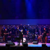 A man singing on a large stage with large band and choir behind him.