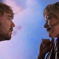 A man looking into the eyes of woman. He seems dishevelled and anxious whereas she seems elegant and confident.
