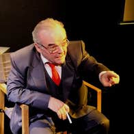 An older man in a suit is seated in a chair, talking and pointing with left finger