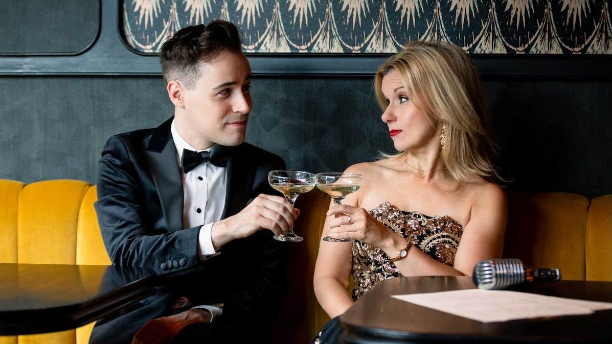 A man and woman in evening attire are seated on a yellow sofa, toasting glasses and looking at each other.