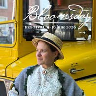 Bloomsday Afternoon Tea Trip, a smiling woman in Edwardian outfit with straw hat standing in front of a vintage yellow bus.