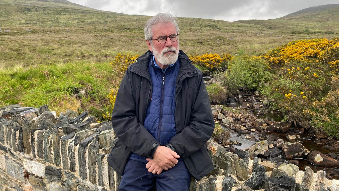 An elderly Gerry Adams, sitting on a wall in the wilderness.