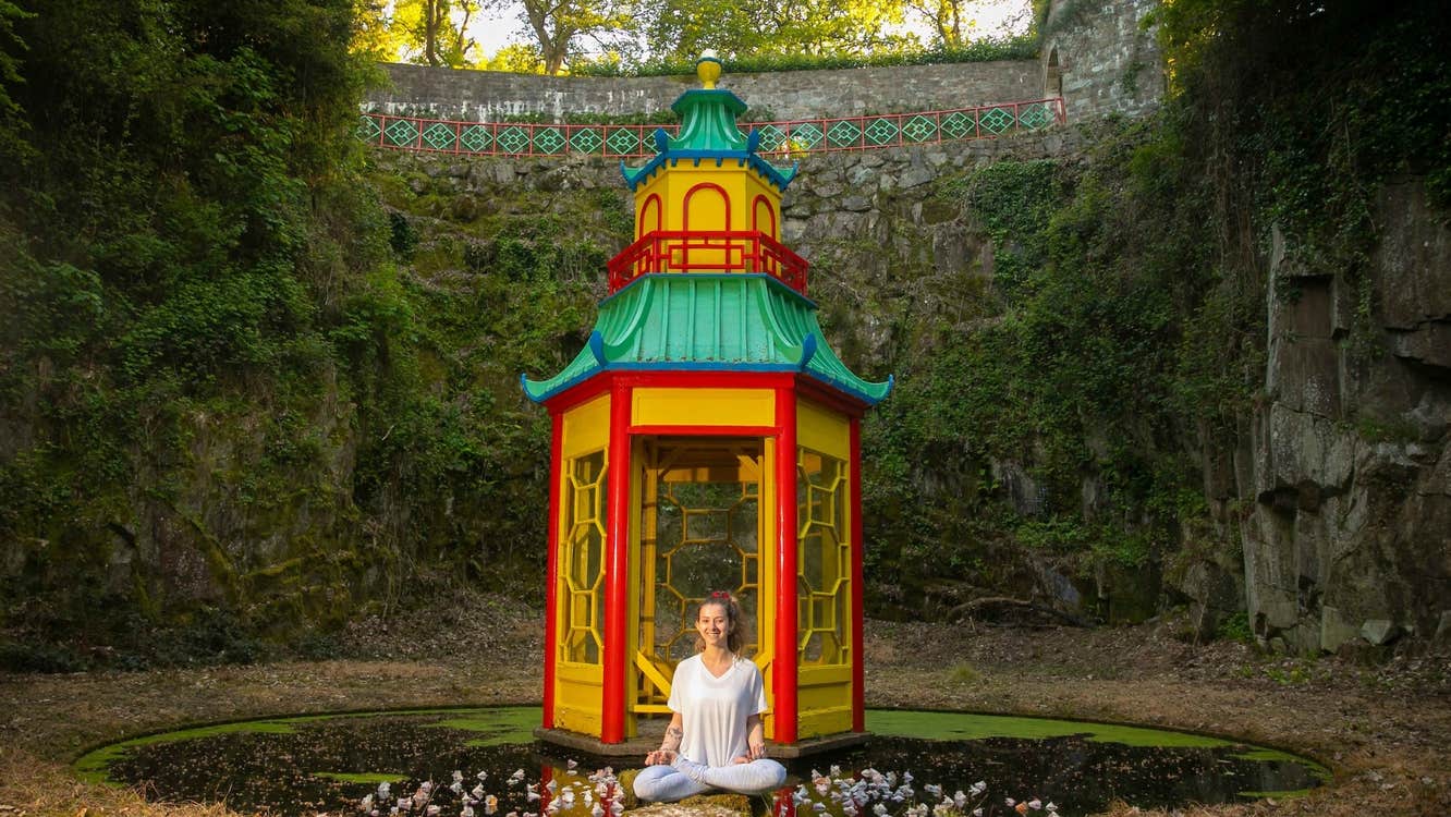 A smiling woman seated crosslegged in front of a small colourful pagoda on a small pond with high green covered walls all around.