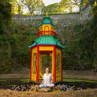 A smiling woman seated crosslegged in front of a small colourful pagoda on a small pond with high green covered walls all around.