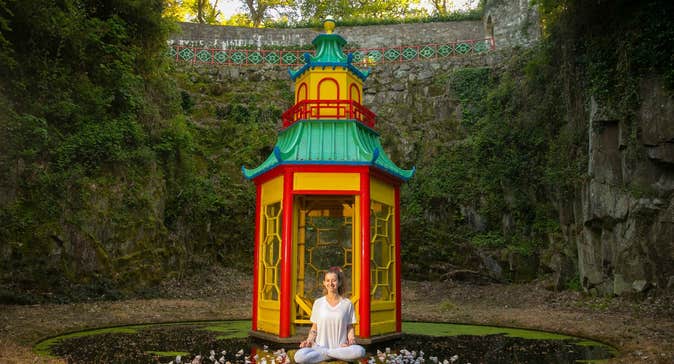A smiling woman seated crosslegged in front of a small colourful pagoda on a small pond with high green covered walls all around.