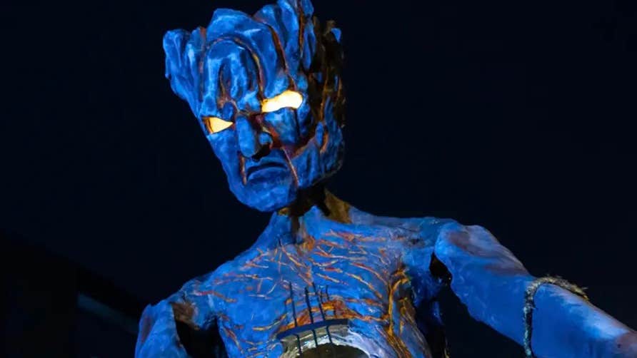 At night, a tall, scary puppet figure in dark blue of person with square head and bright yellow eyes, against black background.