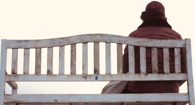 'An tÉalú' Ceoldráma Nua le Seán Óg Ó Duinnín - rear view of person in winter coat and hat seated at one end of a wooden bench.