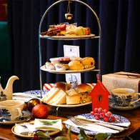 Festive Afternoon Tea stand displayed on a table in the Old Music Shop Restaurant, featuring scones, sandwiches, brioche buns, mince pies, miniature desserts, cups of tea, and a gift box, surrounded by Christmas decorations and ornaments.