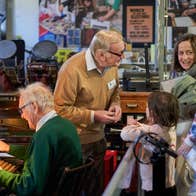 2 older men talking and demonstration old equipment to a small group of adults and children.