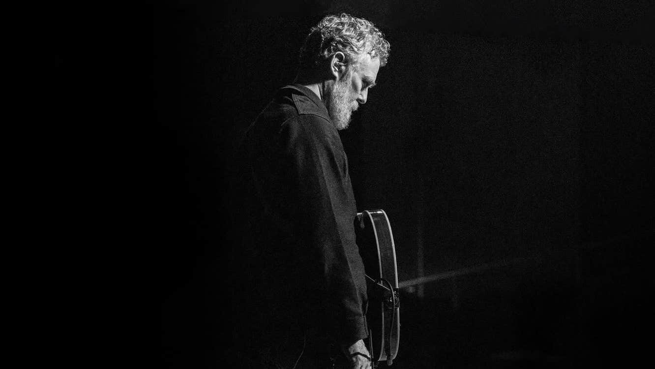 A dimly lit photo of man on a stage with a guitar, looking down.