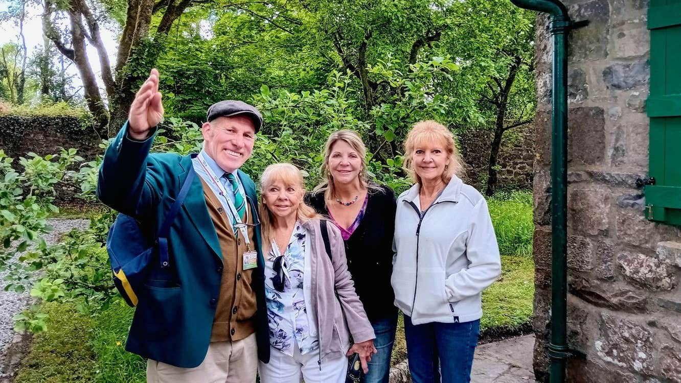 Three people standing with a tour guide next to a stone building with trees in the background