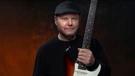 Christopher Cross, a man in a flat cap is holding upright an electric guitar.