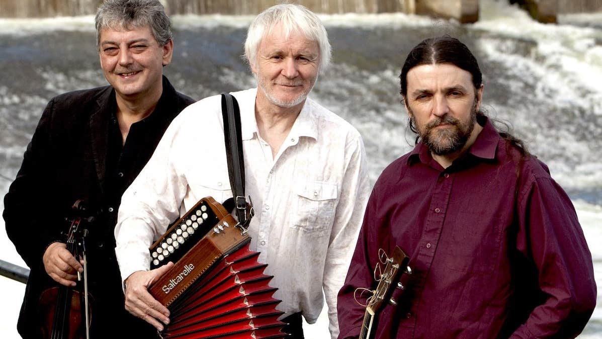 Music Network presents Máirtín O’ Connor Trio, 3 men holding musical instruments standing against railings with cascading water behind them.