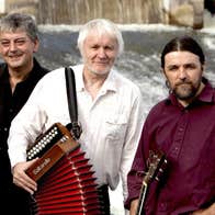 Music Network presents Máirtín O’ Connor Trio, 3 men holding musical instruments standing against railings with cascading water behind them.
