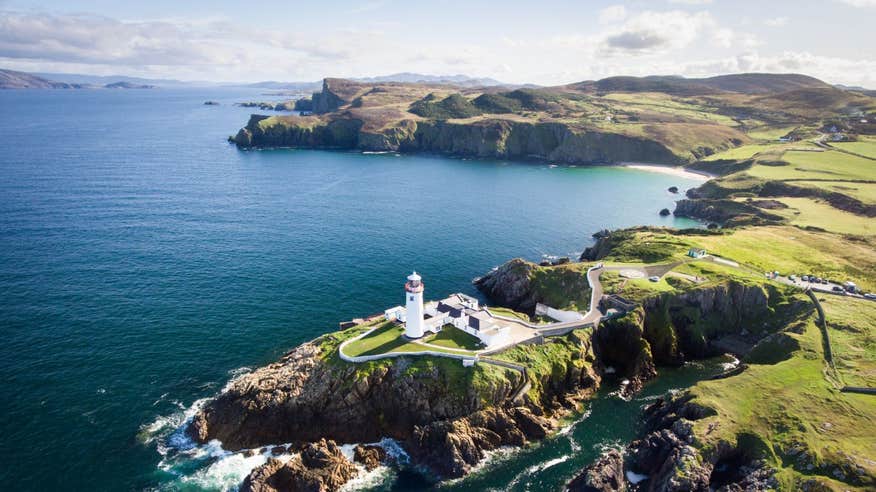 A lighthouse perched on a cliff with a backdrop of green fields and waves crashing against rocks