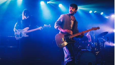 Cian Godfrey of Somebody’s Child plays guitar on stage, with drums visible behind him and atmospheric lights creating a vibrant backdrop.