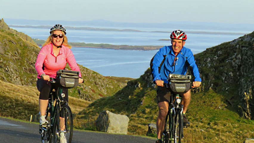 A man and woman cycling up a hill with a view of the sea visible behind them