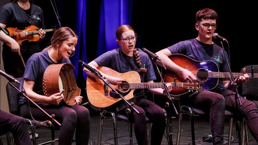 Young adults seated, playing musical instruments, singing as part of a larger group on a stage