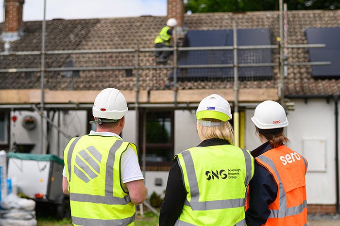 The back of three people wearing hi-vis jackets facing away from the camera towards a house with scaffolding on it