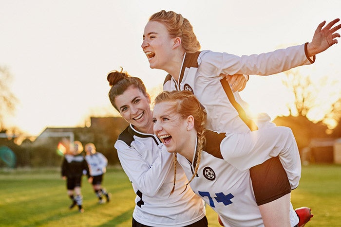 Three young women, in football jerseys. Two are holding the third up. The third has her arms spread out in a pose imitating Rose at the prow of the Titanic. 