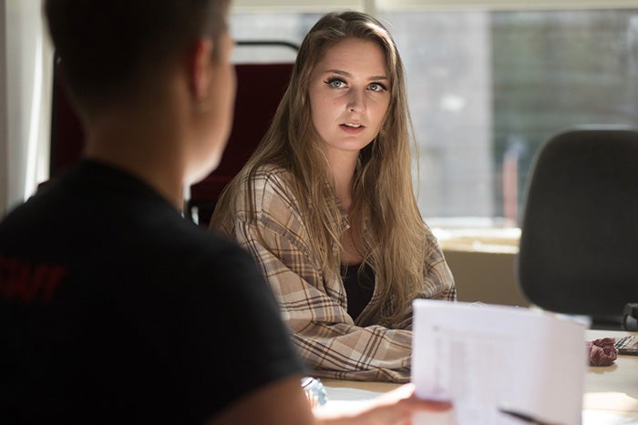 A young woman sat across a table from a young man. The man has some paperwork and appears to be talking to the young woman.